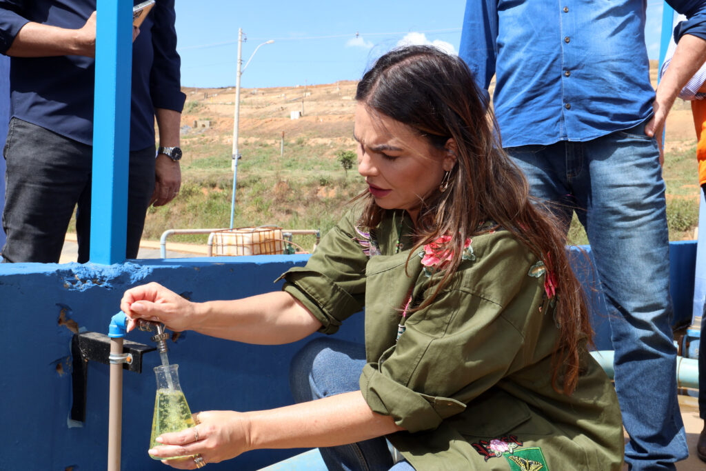 Foto da deputada Paula Belmonte abaixada, abrindo uma torneira de onde sai uma água em tom amarelado.