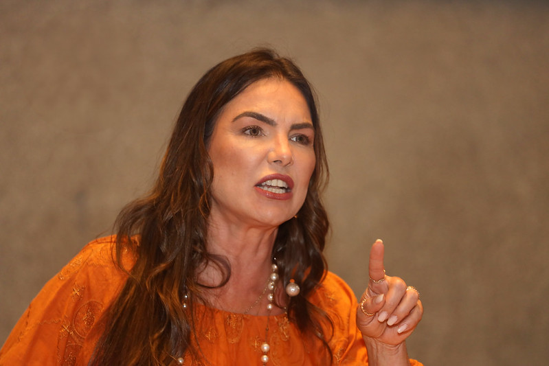 Paula Belmonte veste laranja, com os cabelos soltos e o dedo indicador esquerdo em riste